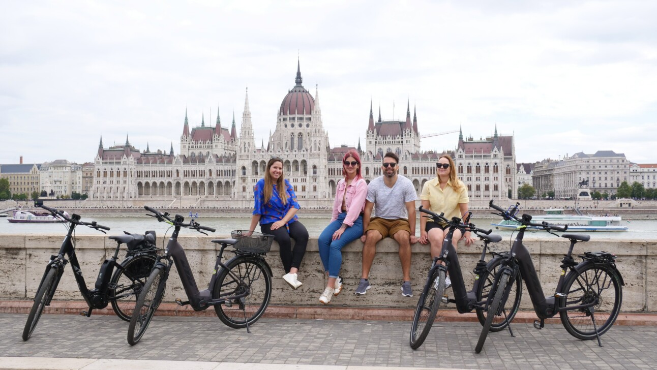 A group of 4 sits along the Danube river with the Hungarian Parliament in the background