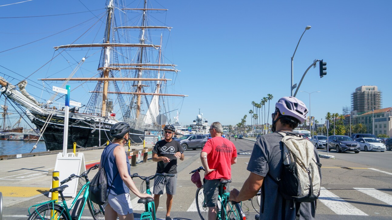 A group of bikers stop along the water where a large sailboat is parked in San Diego