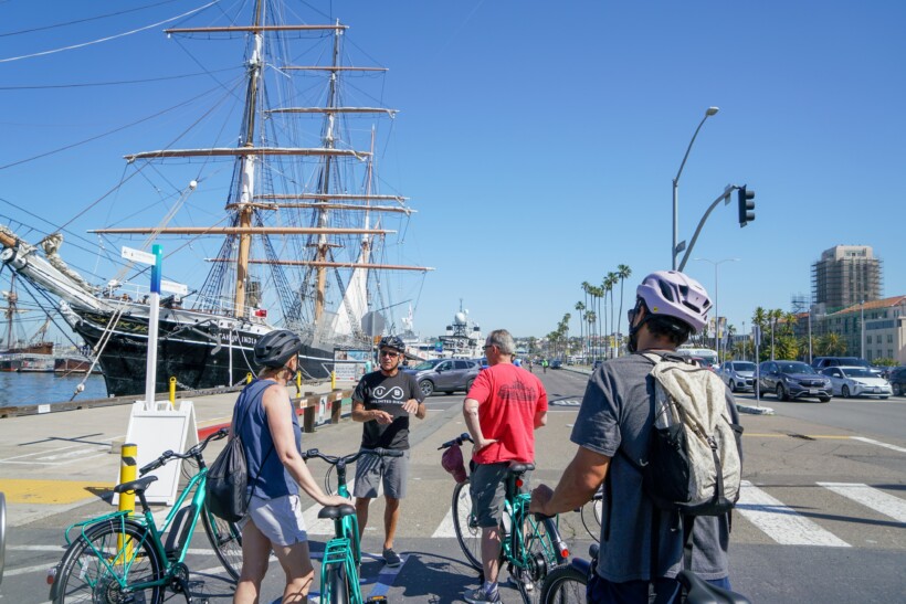 A group of bikers stop along the water where a large sailboat is parked in San Diego