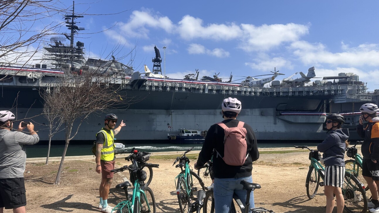 A group of cyclists stop along the water to look at a large military vessel in San Diego