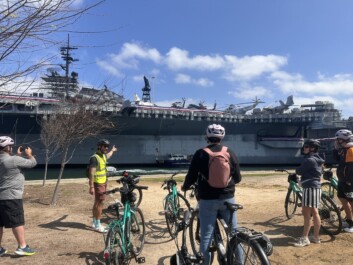 A group of cyclists stop along the water to look at a large military vessel in San Diego