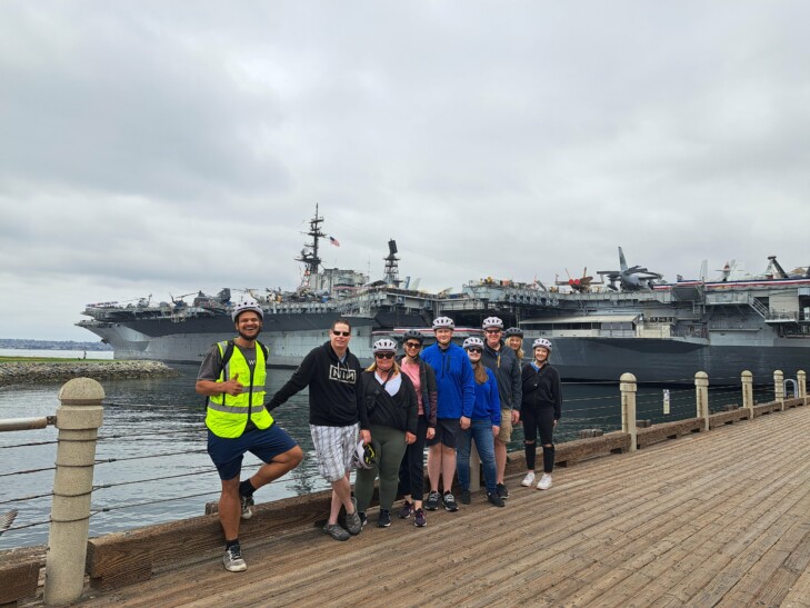 A group of cyclists pose for a photo in front of a large ship docked in San Diego