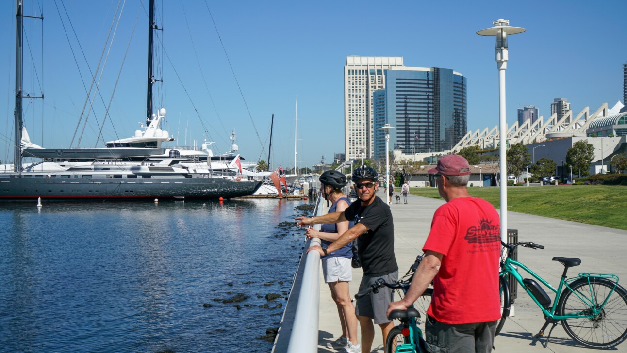 A group of cyclists stop along the water to look at the ships in San Diego