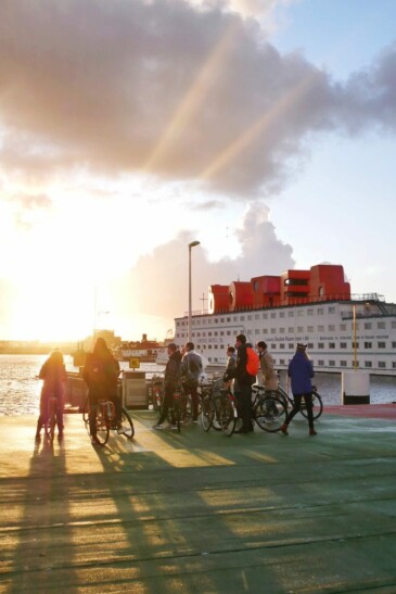 A group of cyclists stand at the end of a canal in Amsterdam as the sun shines through the clouds