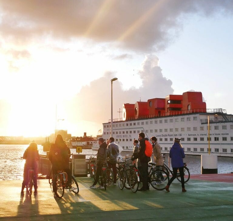 A group of cyclists stand at the end of a canal in Amsterdam as the sun shines through the clouds